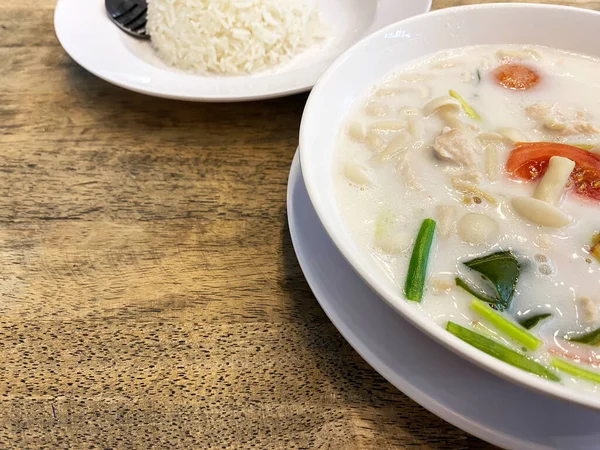 Hindistan cevizi Tom çorbası, ahşap bir masada haşlanmış pirinç ile birlikte beyaz bir kasede taze otlar ve domatesler içerir.