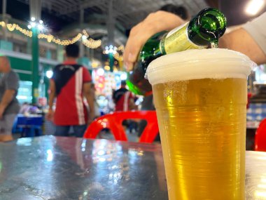 Bira, metalik bir masada açık plastik bir bardağa dökülüyor, bulanık insanlar ve arka plandaki dekoratif ışıklar açık hava pazarında görülebiliyor.