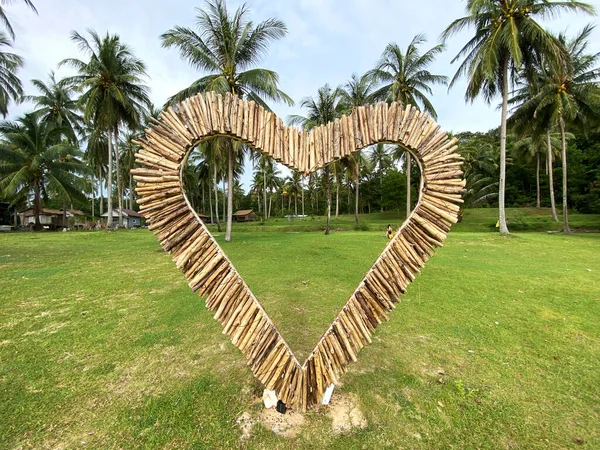 Tahta çubuklardan yapılmış kalp şeklindeki yapı yeşil çimlerin üzerinde duruyor. Arka planda tropik bir ada ortamında palmiye ağaçları var.