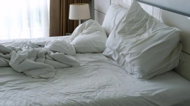 Kırışık beyaz çarşaflar ve yastıklarla yapılmamış yatak düzensizliğin kademeli bir şekilde ilerlediğini ve yatağın kenarında koyu renk bir giysi sergiliyor.