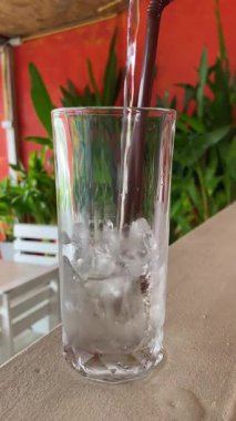 Su içeri akarken buzlu temiz cam, açık havada yemyeşil bitkilerle çevrili kahverengi pipet görünür.