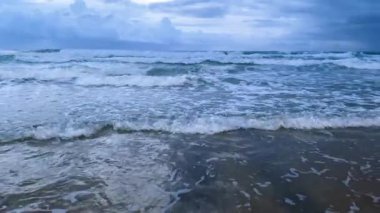 Okyanus dalgaları kıyıya vurur, kumlu sahilde köpük ve dalgalanmalar yaratır, bulutlu bir gökyüzü alacakaranlığın yumuşak ışığını yansıtır.