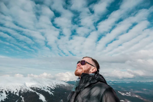 Kışlık ceketli ve güneş gözlüklü sakallı bir adam yukarı bakıyor, karlı bir dağ sırasının tepesinde, güneşte sırılsıklam. Gökyüzü eşsiz, dalgalı altokümülüs bulutlarıyla dolu..