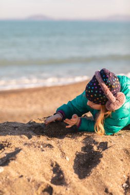 Girl wearing a bright turquoise puffer jacket, a patterned beanie, and pink earmuffs lies on a sandy beach, focused on playing with the sand. In the background, the soft blue waves of the ocean meet a clear sky under the warm glow of the golden hour.