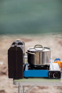 Kapaklı paslanmaz çelik bir tencere katlanabilir bir kamp masasındaki portatif mavi bütan gaz sobasının üzerinde duruyor. Kurulum açık havada, muhtemelen bir kamp alanında ya da suya yakın, yumuşak, odaksız doğal bir arka planda..