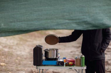 Yüksek açılı, orta ölçekli bir çekim, kamp alanında yemek pişiren siyah ceketli birini yakalar. Yüzü büyük yeşil bir ağ örtüsüyle örtülmüş olan birey, dumanı tüten paslanmaz çelik bir tencerenin üzerinde metal bir kapak tutuyor..