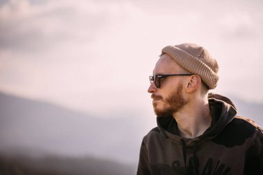 Bereli ve güneş gözlüklü düşünceli sakallı adam dağ manzarasına bakıyor.