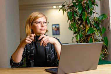 Kadın, dizüstü bilgisayarının yanında kahve fincanı tutarak yansıtıcı bir mola veriyor..