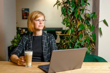 Bir kadın rahat bir ortamda dizüstü bilgisayarıyla çalışırken kahve içer..