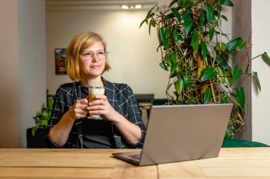 Gülen kadın elinde dizüstü bilgisayarla tahta masada otururken bir bardak kahve kaldırıyor..