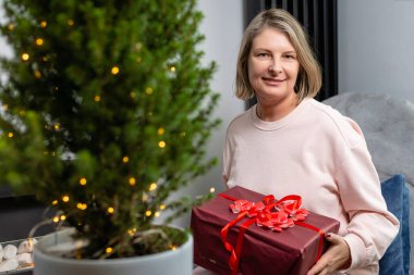 Bayram hediyesini tutarken süslü ağacın yanında poz veren rahat bir kadın..