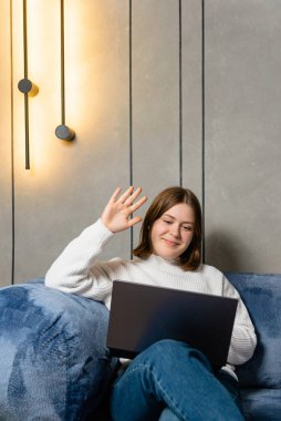 Oturma odasındaki mavi koltukta oturan mutlu genç bir kadın aktif olarak bir video konferans görüşmesi yapıyor ve kameraya el sallıyor..