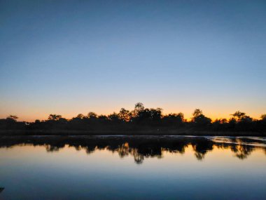 Sakin bir göl üzerinde sakin bir günbatımı sahnesi. Durgun suya yansıyan siyah siluetler. Gökyüzü, ufkun yakınındaki ılık portakaldan yukarıdaki derin mavime doğru yumuşak bir eğim gösteriyor. Rahatlamaya uygun doğal bir manzara yaratıyor..