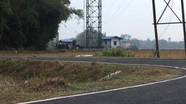 Arka planda elektrik iletim kuleleri ve küçük kamu hizmetleri binaları olan açık tarım arazisinden geçen sakin kırsal yol. Kırsal altyapı, güç dağılımı ve açık tarım manzarasını gösteren barışçıl kırsal bölge ortamı.