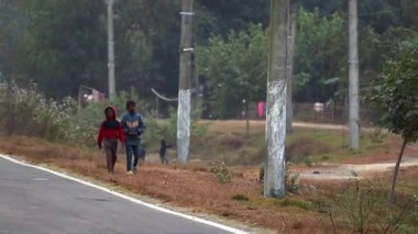 Assam, Hindistan Ocak 2026: Kırsal bir köyde elektrik direkleri, ağaçlar ve tarlalarla kaplı asfalt bir yolda yürüyen iki çocuk. Sahne, günlük kırsal yaşamı, çocukların günlük hareketlerini, köyün çevresini temsil ediyor..