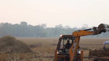 Dibrugarh, Assam, Hindistan, 10 Ocak 2026: Bir kazıcı Assam 'daki arazi çalışmaları sırasında işçiler makineleri çalıştırırken açık bir kırsal alanda bir traktör römorkuna toprak yüklüyor. Sahne gerçek tarım ve inşaat işçilerini gösteriyor..