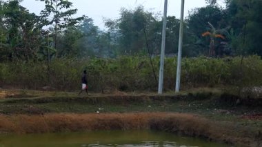 Dibrugarh, Assam, Hindistan, 15 Ocak 2026: Ağaçlar ve bitki örtüsüyle çevrili kırsal bir göletin yanında duran bir adam, arka planda elektrik direkleriyle, Hindistan 'ın kuzeydoğusundaki günlük köy yaşamını ve kırsal çevreyi tasvir ediyor..