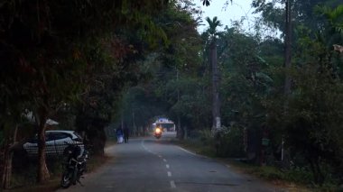 Dibrugarh, Assam, Hindistan, 15 Ocak 2026: Yoğun ağaçların altında bir motosiklet yaklaşırken dar bir yolda yürüyen yayaları gösteren sakin bir akşam sokak sahnesi. Görüntü, yol kenarındaki bitki örtüsüyle küçük bir Hint kasabasında günlük yaşamı yakalıyor..