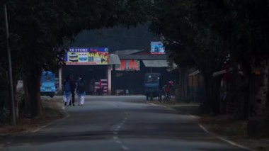 Dibrugarh, Assam, Hindistan, 15 Ocak 2026: Yayalar, Dibrugarh 'taki küçük işletmelerin ve yerel dükkanların yakınlarındaki sık ağaçların gölgelediği sessiz bir yol boyunca yürüyor. Sahne, yerel taşımacılığın yer aldığı küçük bir Hint kasabasındaki günlük yaşamı yansıtıyor..
