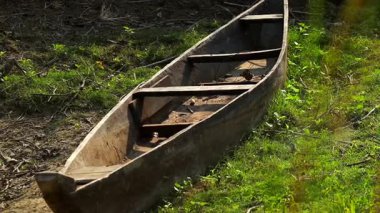 Geleneksel ahşap bir kanonun yakın plan görüntüsü çimenli bir nehir kıyısında dinleniyor, eskimiş ahşap dokular ve basit el yapımı yapılar sergiliyor. Doğal güneş ışığı kırsal detayları vurgular, sakin bir kırsal ortam yaratır..