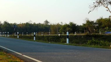 Yeşil bitki örtüsü ve siyah-beyaz yol işaretleri arasında sessiz bir asfalt yol uzanıyor. Ağaçlar ve ormanlık alanlar arka plana doğru uzanıyor ve sakin bir kırsal atmosfer yaratıyor. Sahne kırsal ulaşımı temsil ediyor.