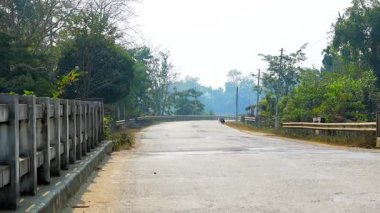 Ağaçlar, yol kenarındaki bariyerler ve yumuşak sabah sisi ile çevrili yeşil köy manzarası boyunca yavaşça kıvrılan huzurlu kırsal yol. Boş asfalt yol seyahat, ulaşım ve doğa temaları için ideal sakin bir kırsal atmosfer yaratır..