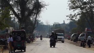 Dibrugarh, Assam, Hindistan, 12 Şubat 2026: Bir motosikletli, küçük dükkanlar, park edilmiş araçlar ve otomobillerle dolu işlek bir yarı-şehir yolu boyunca ilerliyor. Hafif trafik bulutlu bir gökyüzü altında ilerlerken, bölge sakinleri yol kenarında yürüyor.