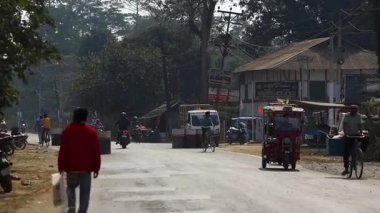 Dibrugarh, Assam, Hindistan, 12 Şubat 2026: Motosikletler, bisikletler ve otomobiller de dahil olmak üzere karışık trafik, küçük dükkanlar ve elektrik direkleriyle kaplı yarı kentsel yol kenarında ilerliyor. Yayalar günlük yaşam olarak yol kenarında yürürler.