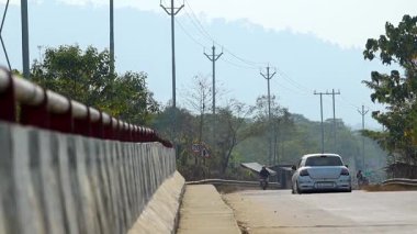 Dibrugarh, Assam, Hindistan, 12 Şubat 2026: Beton bariyerler ve yol kenarındaki elektrik direkleriyle çevrili kırsal bir otoyol boyunca beyaz bir araba sürüyor. Arka planda sisli tepeler ve ağaçlarla kırsal yolda hafif trafik hareket ediyor.