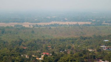 Puslu bir gökyüzünün altında dağınık evler, ağaçlar ve tarım arazileri olan yemyeşil bir vadinin geniş panoramik manzarası. Manzara yoğun bitki örtüsü ve açık tarım arazisiyle çevrili huzurlu bir kırsal yerleşim gösteriyor.