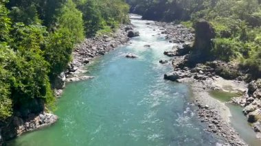 Dağlık tepelerle çevrili kayalık kıyılar ve yoğun yeşil ormanlar arasında kıvrılan turkuaz bir nehrin çarpıcı geniş manzarası. Güneş ışığı, berrak akan suyu ve yemyeşil bitki örtüsünü aydınlatır. Huzurlu ve ferahlatıcı bir doğal manzara yaratır..