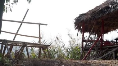 Yüksek sazlı bir barınağın yanında yüksek otlarla çevrili kırsal bambu oturma alanı. El yapımı ahşap yapılar geleneksel işçiliği yansıtıyor ve kırsal alanda basit bir dinlenme alanı sağlıyor..
