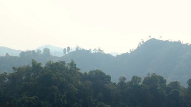 Yuvarlanan tepeleri kaplayan yoğun yeşil orman yumuşak sise dönüşüyor. Katmanlı dağ manzarası derinlik ve sakin bir atmosfer yaratır, huzurlu bir kırsal çevrenin doğal güzelliğini yakalar..