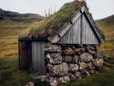 Geleneksel bir çim çatılı ev Faroe Adaları 'nın manzaralı bir bölgesinde duruyor. Yeşil tepeler ve kayalık arazilerle çevrili bu yapı bulutlu bir gökyüzü altında yerel mimariyi sergiliyor..