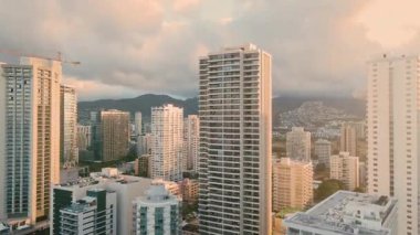 Güneş batarken şehrin yüksek binalarının manzarası. Gökyüzü bulutları ve yumuşak ışığı gösteriyor. Şehir manzarası, uzaktaki yüksek yapıları ve çevresindeki tepeleri kapsar..