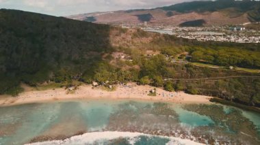 İnsanlar Hawaii 'de güneşli bir günün tadını çıkarıyorlar. Su berrak ve kum sıcak. Bölgeyi çevreleyen tepeler ve ağaçlar ziyaretçiler için güzel bir manzara oluşturuyor..