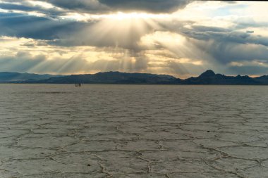 Güneş ışığı bulutları delip geçerek kuru ve çatlak yüzeyi olan bir tuzu aydınlatıyor. İnsanlar gün batımında çölde olay çıkararak uzaklarda yürür..