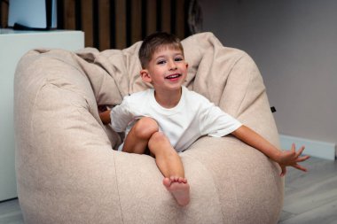 Geniş Fasulye Torbada Rahatlayan Çocuk, Gülümseyen Geniş, Rahat Tişört, Konforlu Salon Sahnesi, Yaslanan Duruş, Oyuncu Ulaşım, Sıcak Ortam Işığı, Kaygısız Rahatlık, Ev Rahatlığı