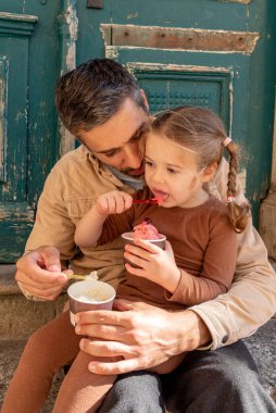 Aile babası ve küçük kızı dondurma yer, baba ve kızı soğuk meyve tatlısı yer.