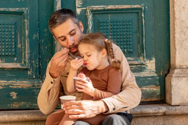 Aile babası ve küçük kızı dondurma yer, baba ve kızı soğuk meyve tatlısı yer.