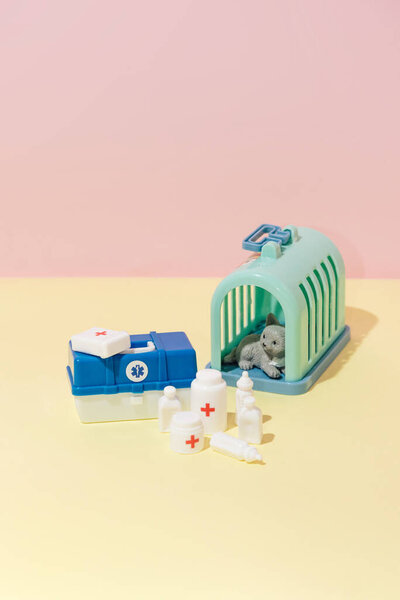Toy medical suitcase with veterinary medicines and instruments and cat cage on pink and yellow background creative composition