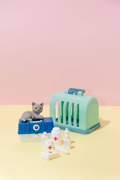 Toy medical suitcase with veterinary medicines and instruments and cat cage on pink and yellow background creative composition