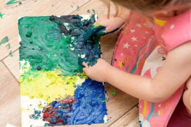 Küçük tatlı kız yerde otururken parlak renkli boyalarla el çiziyor.