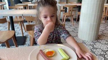 Kahvaltıda omlet, salatalık ve sosis yiyen tatlı bir kız çocuğu.