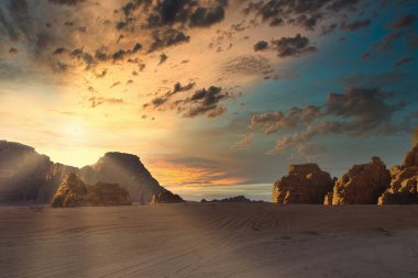 Bulutlar gökyüzünde toplanırken güneş dağların ardında batıyor. Kumlu zemin görülebilir, kayalık oluşumları vurguluyor. Wadi Rum Çölü, Ürdün, Ay Vadisi