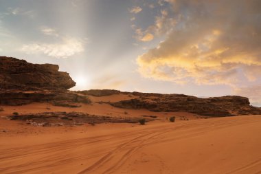 Güneş kırmızı bir çöl manzarasının üzerinde batar. Kum arazisi, kayalık oluşumlarla desteklenen bir şekilde zemin boyunca uzanır. Işık bulutların arasında parlıyor, alacakaranlıkta güzel bir manzara yaratıyor. Wadi Rum, Ürdün.
