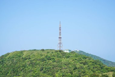 Telekomünikasyon kulesi açık mavi gökyüzünün altında yemyeşil bir dağın zirvesinde..