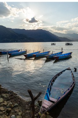 Pokhara, Nepal 'deki Phewa Gölü' nde (Phewa Tal) geleneksel ahşap kayıklar, günbatımında Himalaya eteklerini ve akşam gökyüzünü yansıtan sakin suyla gölün kıyısında demirlemiş renkli kayıklar..