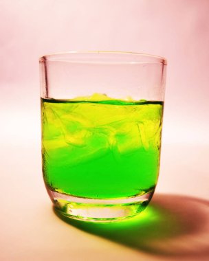 Parlak yeşil sıvı ve buz küpleriyle dolu, parlak pembe ve sarı gradyan arkaplan üzerine kurulmuş parlak bir bardağın görsel olarak çarpıcı bir görüntüsü..