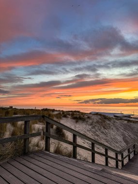 Kumlu kum tepeleri ve kıyıya uzanan ahşap bir geçitle sakin denizin üzerinde inanılmaz bir gün batımı. Sıcak akşam ışığı ve renkli gökyüzü huzurlu bir yaz ortamı yaratır..